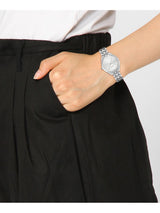 A close-up of a person wearing a silver analog watch with a matching metal link bracelet, paired with black pleated trousers and a white top.