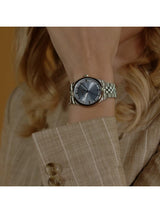 A silver-toned women's watch with a textured sunburst dial and matching metal link bracelet, shown worn on a woman's wrist. The watch features a round case and a silver-tone index bezel.