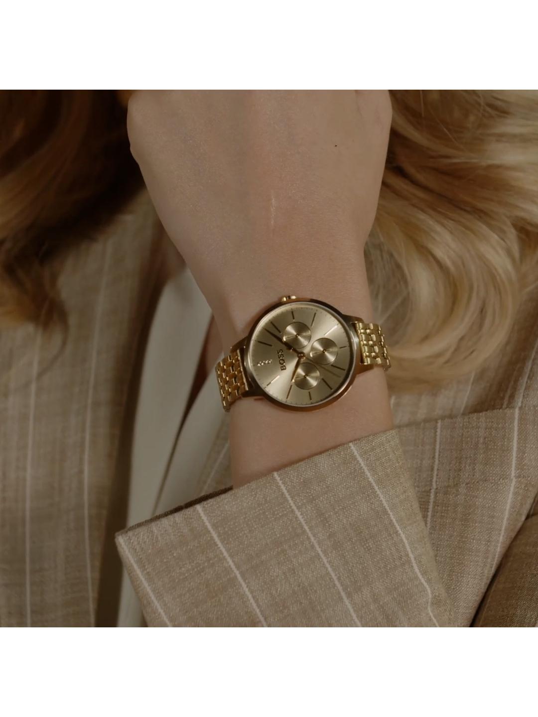 A close-up of a woman's wrist wearing a gold-tone Michael Kors watch with a matching metal link bracelet, styled with a beige pinstripe blazer.