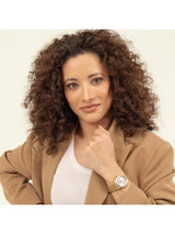A woman with voluminous curly brown hair is shown from the chest up, wearing a tan blazer over a white top and a gold-tone watch with a white dial on her wrist, set against a plain off-white background.