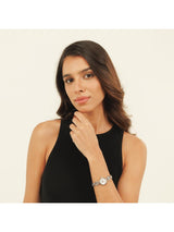 A woman with dark brown hair wearing a black sleeveless top and a silver-toned watch with a white dial, posing against a solid off-white background.