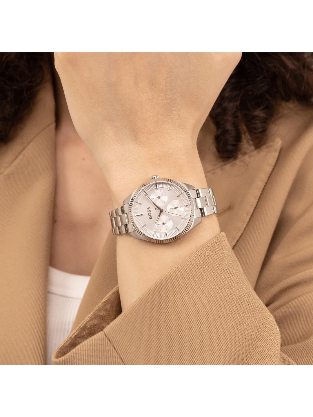 A silver-toned women's analog watch with a matching metal link bracelet, shown worn on a person's wrist. The watch features a textured bezel, a white dial with subtle branding, and is framed by a tan blazer and white top.