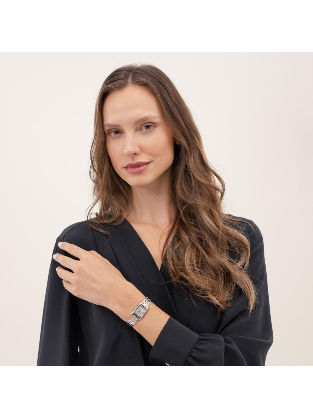A woman with long wavy brown hair is wearing a black long-sleeve blouse and a silver-toned rectangular watch with a clear crystal face, posed against a plain light beige background.