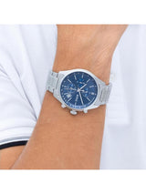 A silver stainless steel wristwatch with a blue dial and date window, shown worn on a person's wrist against a white shirt background.