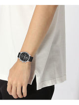 A wristwatch with a black dial and black strap worn on a person's wrist, shown against a white shirt and black pants.
