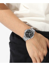 A silver stainless steel wristwatch with a black dial and bezel, worn on a person's wrist. The person is wearing a white top and black trousers, photographed against a clean white background.