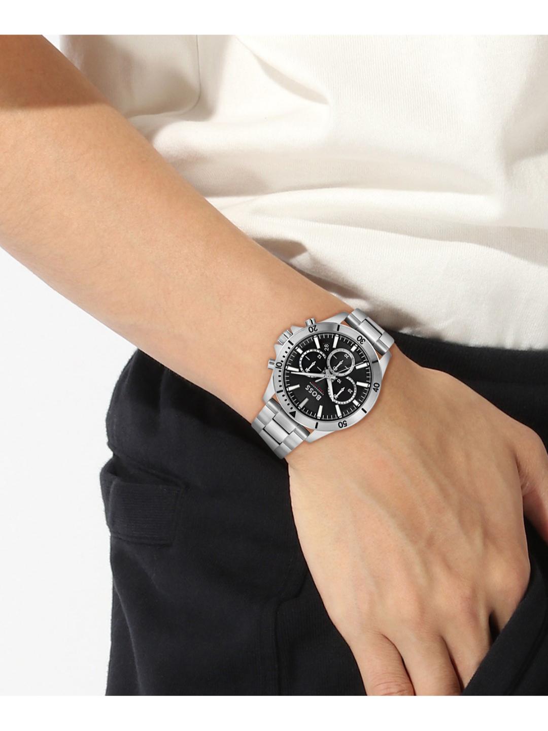 A silver stainless steel wristwatch with a black dial and bezel, worn on a person's wrist. The person is wearing a white top and black trousers, photographed against a clean white background.