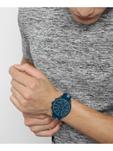 A close-up of a person wearing a blue analog watch with a matching blue strap, shown against a gray heathered t-shirt and a light gray background.