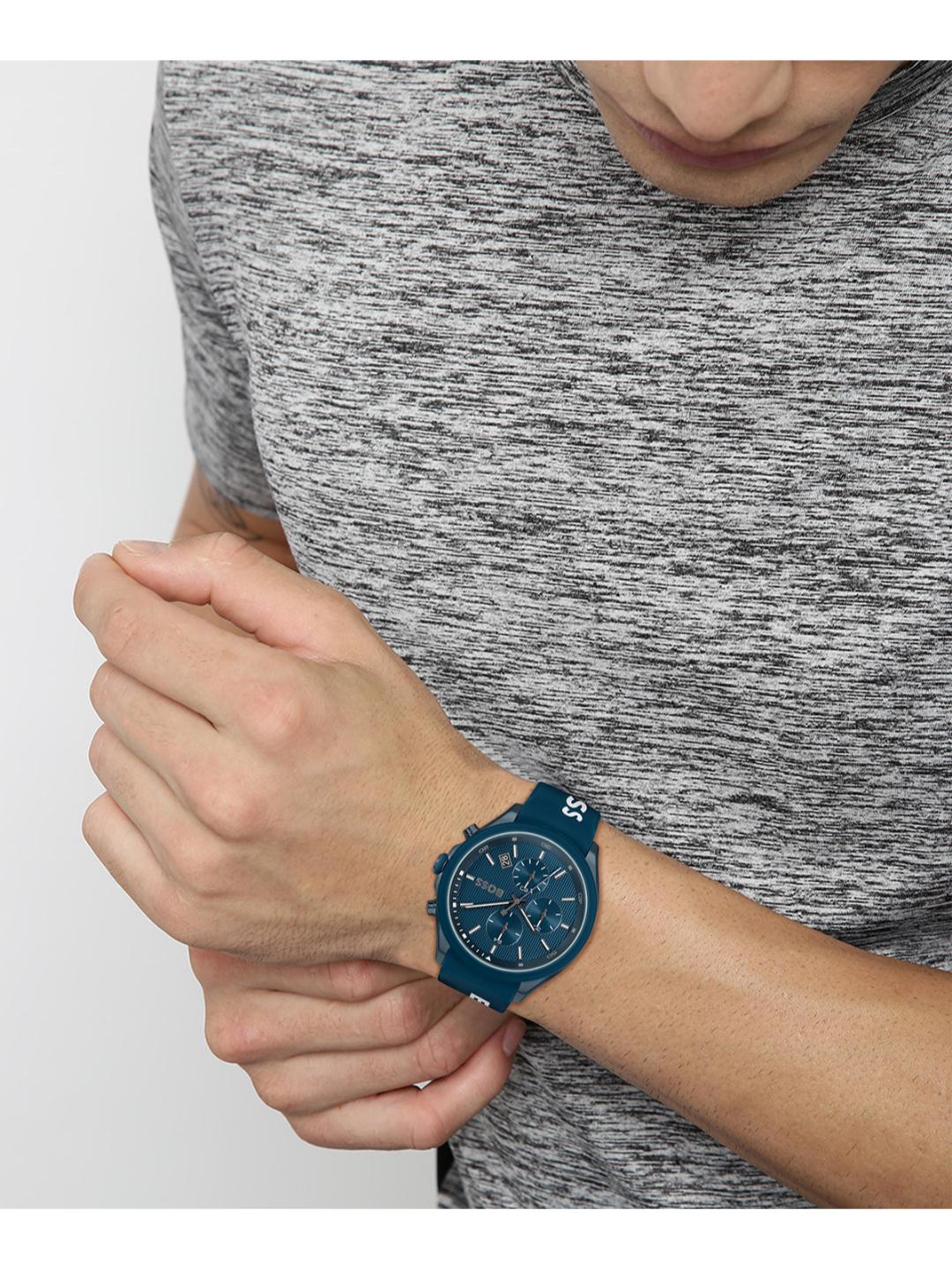 A close-up of a person wearing a blue analog watch with a matching blue strap, shown against a gray heathered t-shirt and a light gray background.