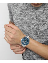 A silver-toned metal watch with a blue dial and bezel, worn on a person's wrist. The person is wearing a gray heathered athletic t-shirt against a light gray background.