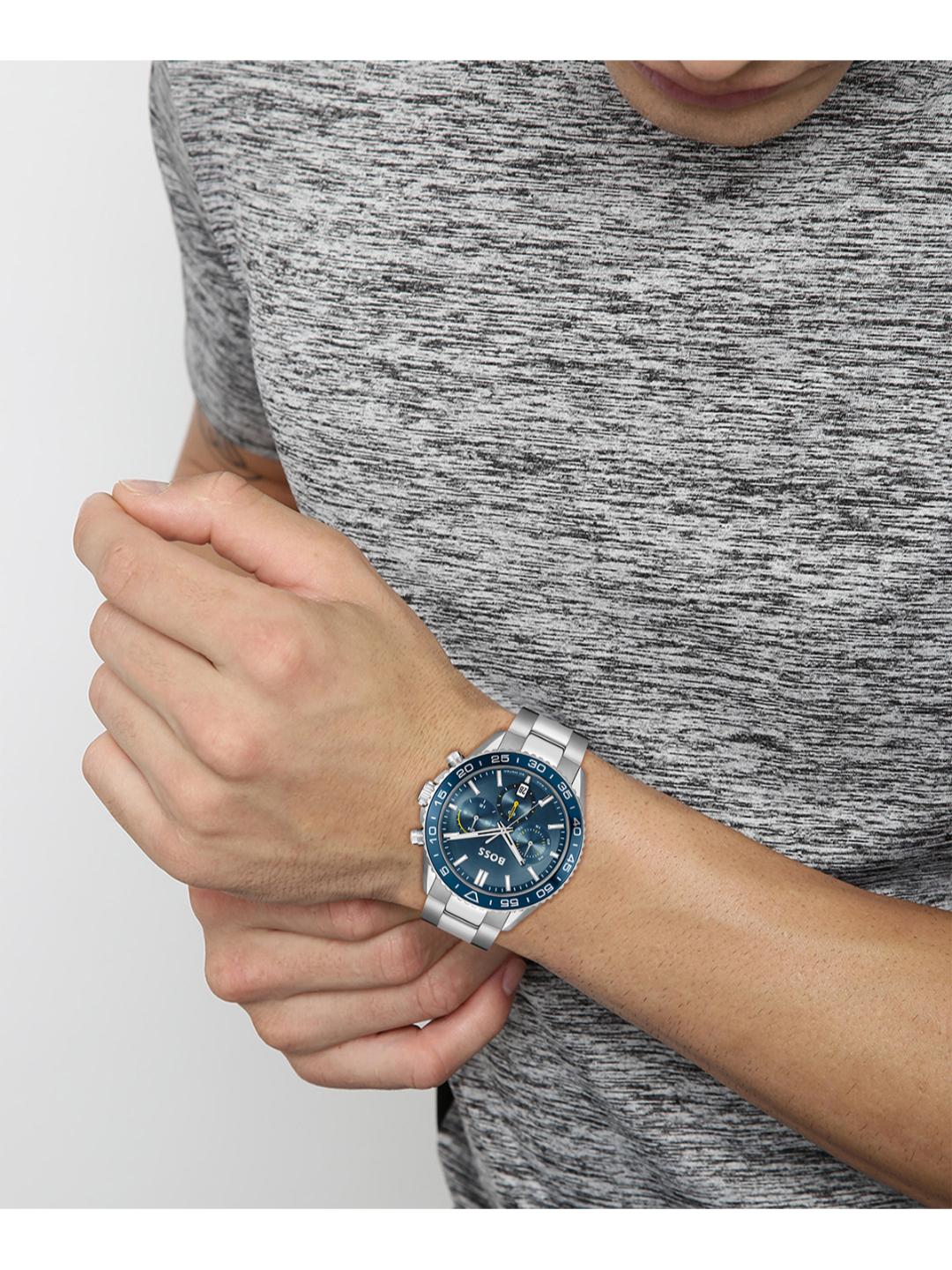 A silver-toned metal watch with a blue dial and bezel, worn on a person's wrist. The person is wearing a gray heathered athletic t-shirt against a light gray background.