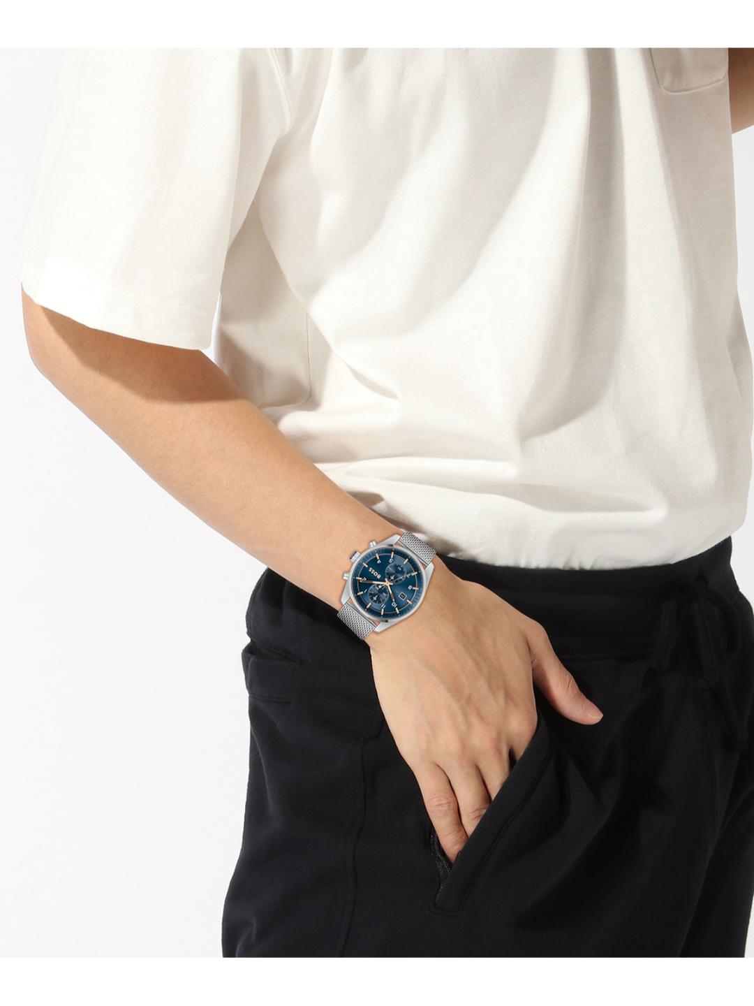 A person wearing a silver-toned wristwatch with a blue dial, styled with a white short-sleeve shirt and black trousers against a plain white background.