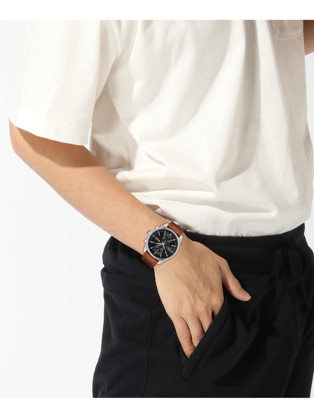 A close-up of a person wearing a brown leather strap watch with a black dial and silver-tone case, paired with a white t-shirt and black pants.