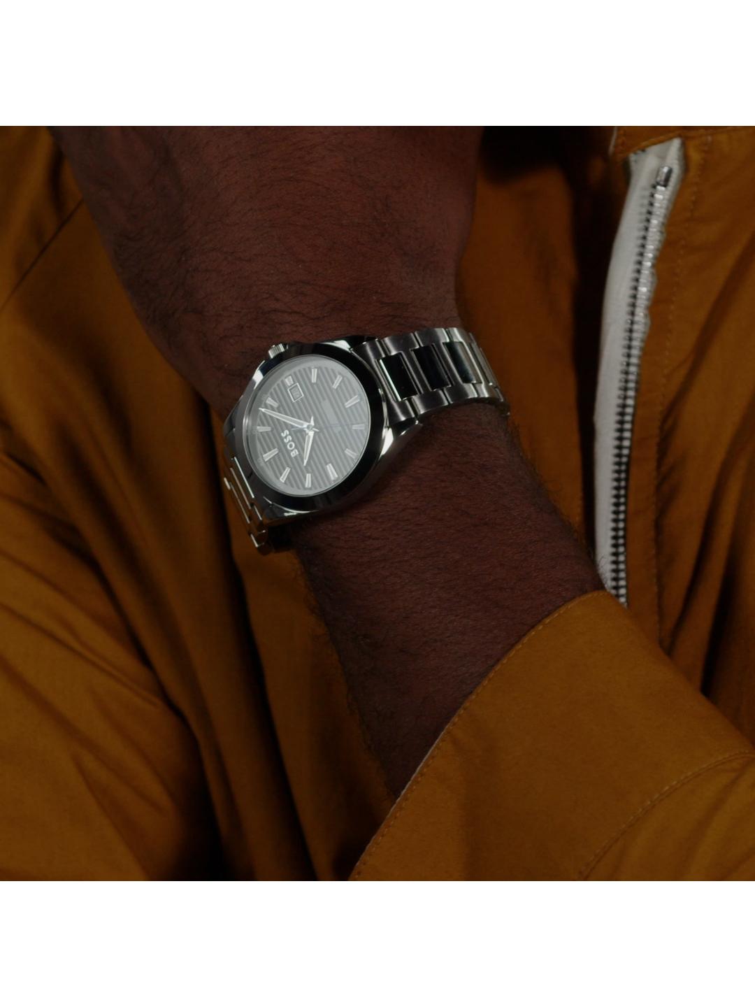 A close-up of a wrist wearing a silver-toned analog watch with a textured dial and metal link bracelet, worn under a mustard yellow zippered jacket.