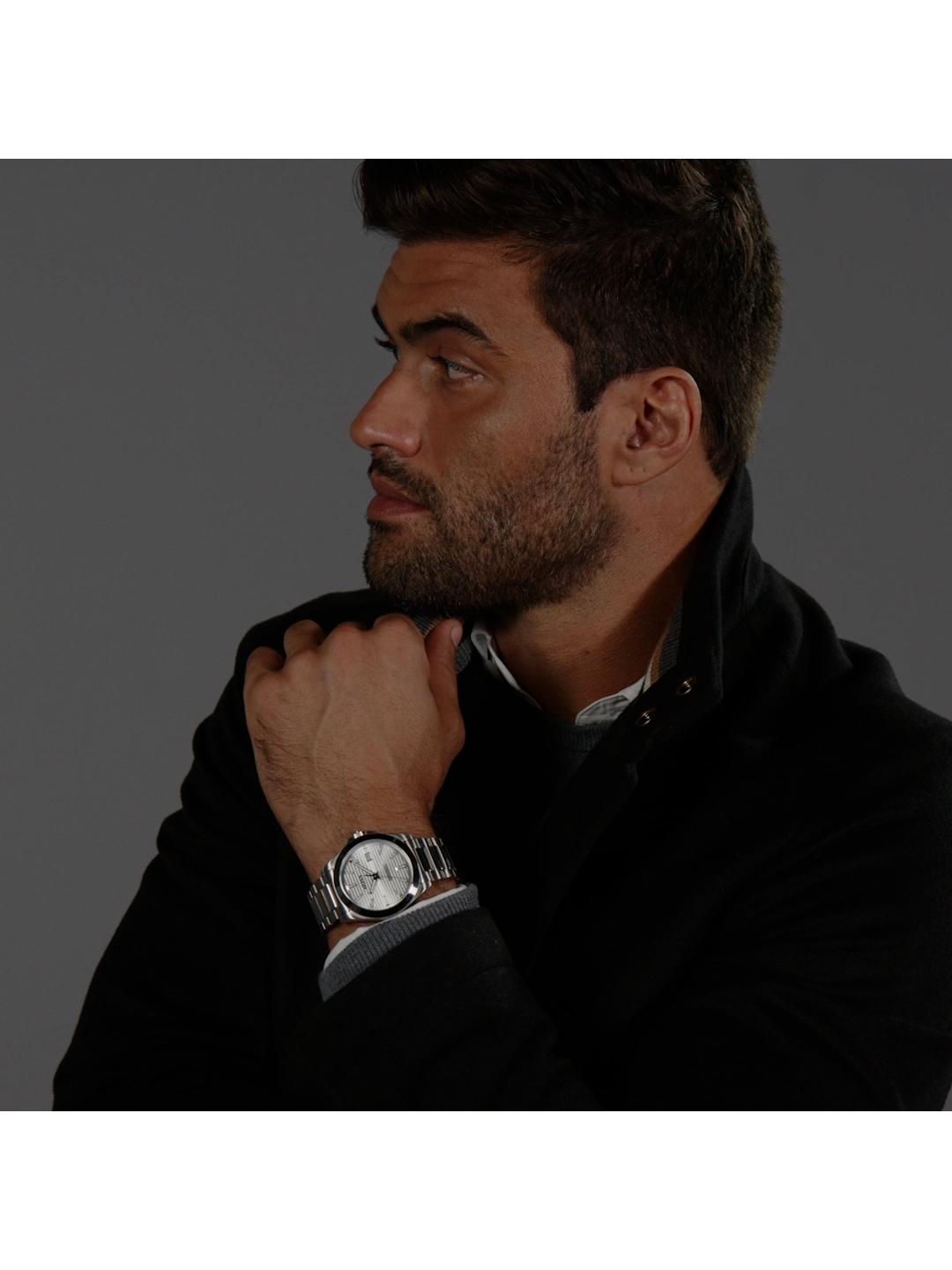 A man in a black jacket wearing a silver-toned wristwatch with a white dial, shown in a close-up profile against a dark gray background.