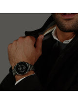 A close-up of a man's hand wearing a silver metal link watch with a black dial, styled with a dark jacket and white shirt.