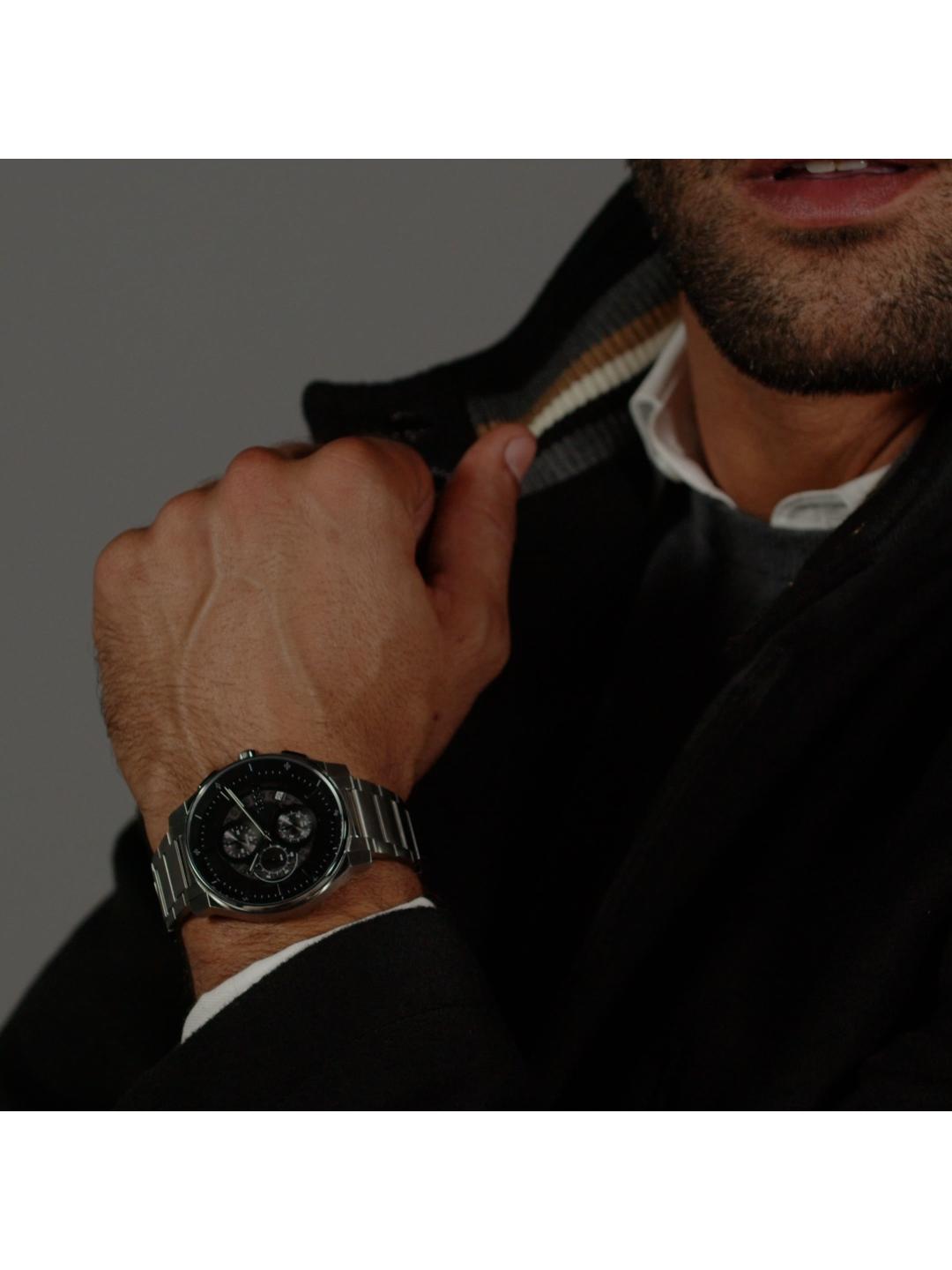 A close-up of a man's hand wearing a silver metal link watch with a black dial, styled with a dark jacket and white shirt.