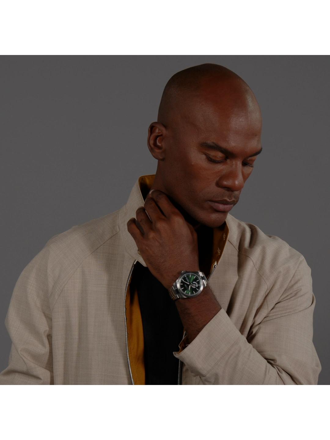 A man wearing a light beige overshirt over a black shirt and gold tie, wearing a silver watch with a green dial on his wrist, set against a dark gray background.