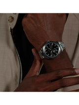 A close-up of a person's wrist wearing a silver-toned metal watch with a black dial and matching black-and-silver bracelet.