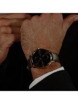 A close-up of a man's wrist wearing a Gucci watch with a black dial, gold-tone case, and dark gray strap, styled with a black suit jacket and white shirt cuff.