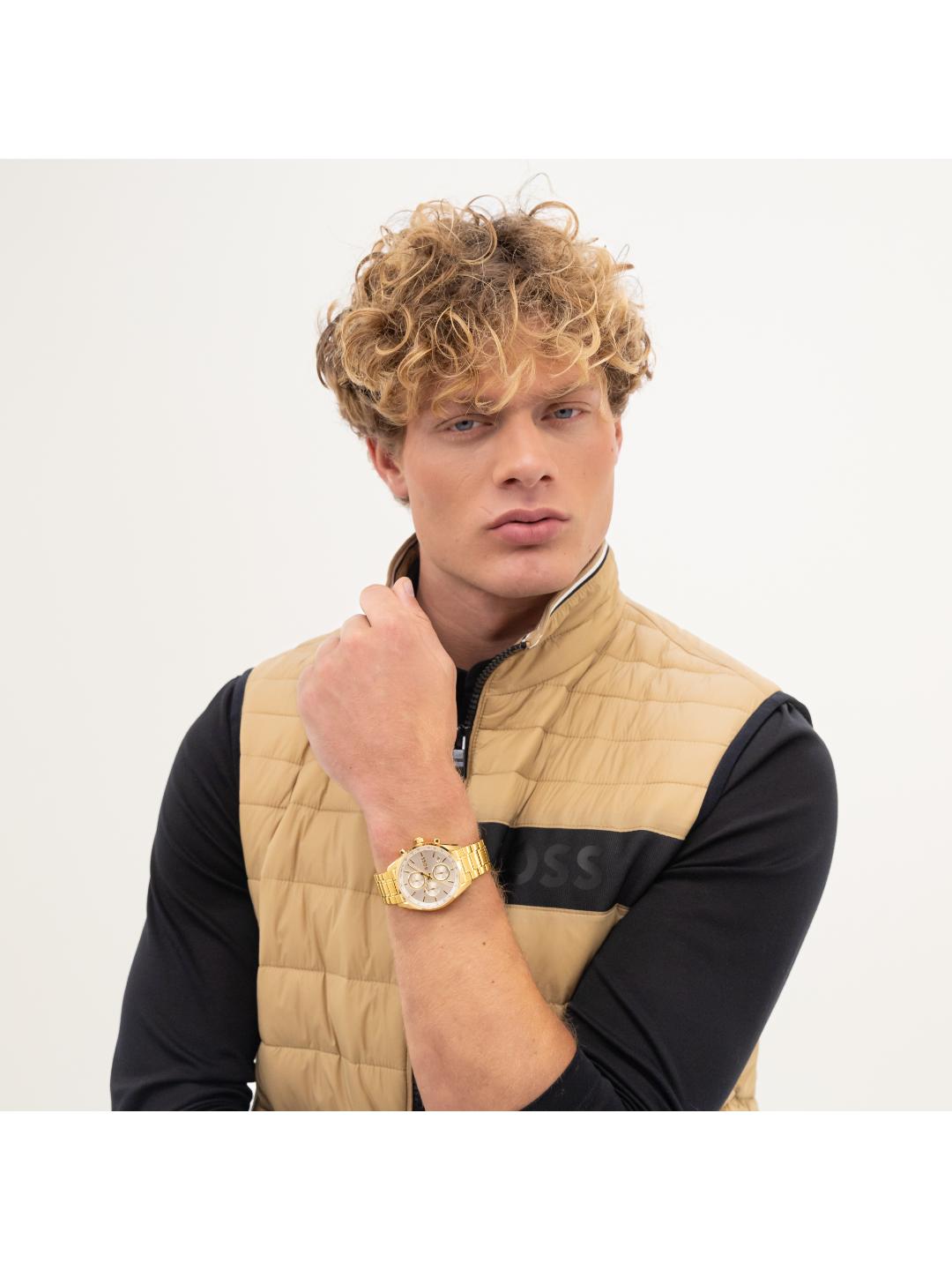 A man with curly blond hair wearing a beige quilted vest over a black long-sleeve shirt, accessorized with a gold watch, posed against a light gray studio background.