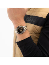 A close-up of a person's wrist wearing a Hugo Boss watch, with a tan and black puffer vest in the background.