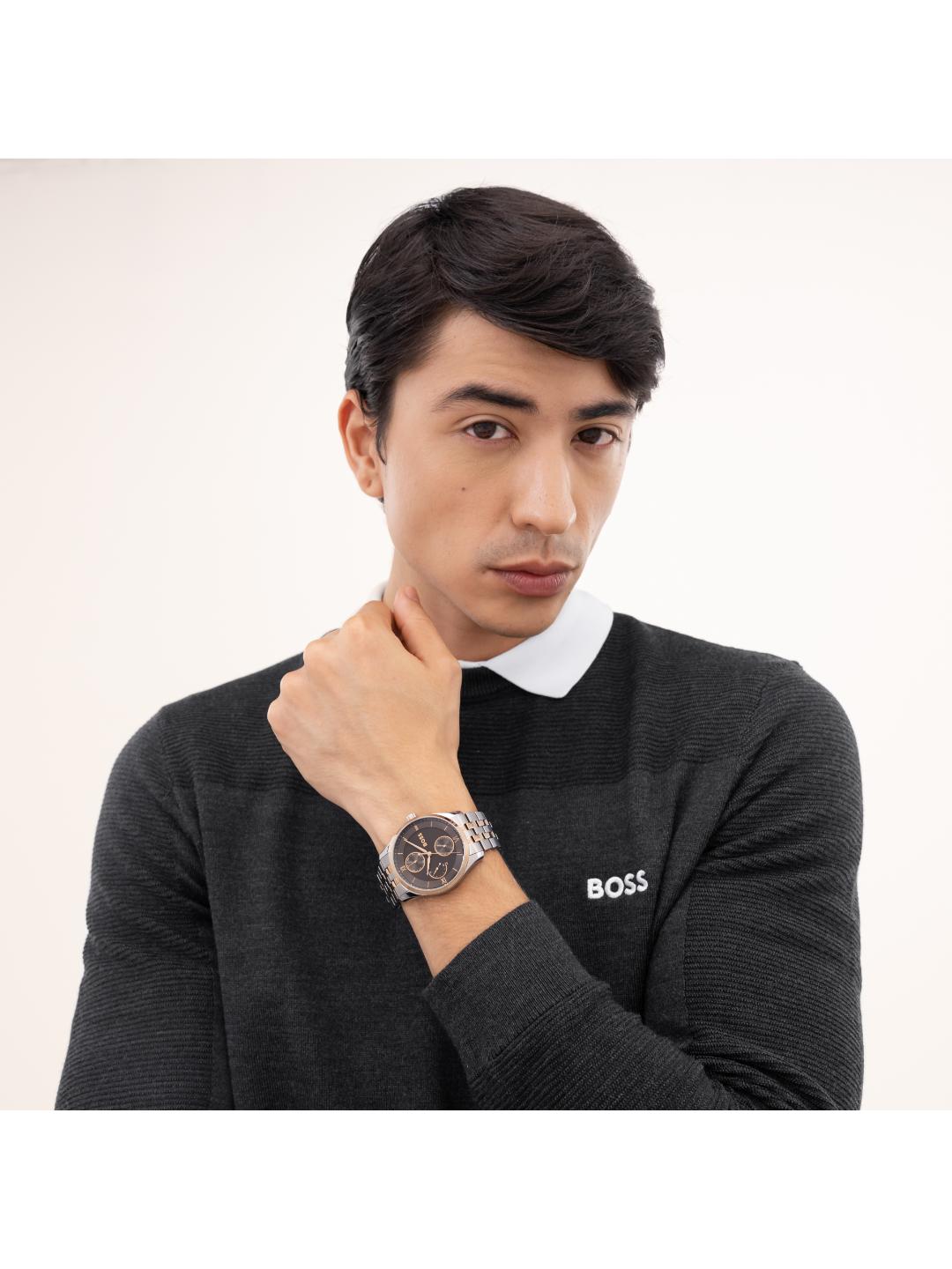 A man wearing a dark charcoal gray BOSS knit sweater over a white collared shirt, wearing a BOSS watch on his wrist. The watch has a dark dial and a silver-toned metal case with a silver link bracelet.