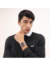 A man wearing a dark charcoal gray BOSS knit sweater over a white collared shirt, wearing a BOSS watch on his wrist. The watch has a dark dial and a silver-toned metal case with a silver link bracelet.