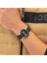 A close-up of a person's wrist wearing a Hugo Boss analog watch with a black dial and black strap, set against a beige puffer jacket with a black logo stripe.