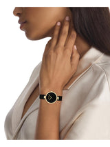 A woman wearing a minimalist wristwatch with a black strap, black dial, and gold-tone case, shown in a close-up side profile against a white background.