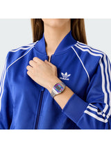 A close-up of a person wearing a royal blue Adidas track jacket with white stripes and a white Adidas logo on the chest, accessorized with a digital watch.