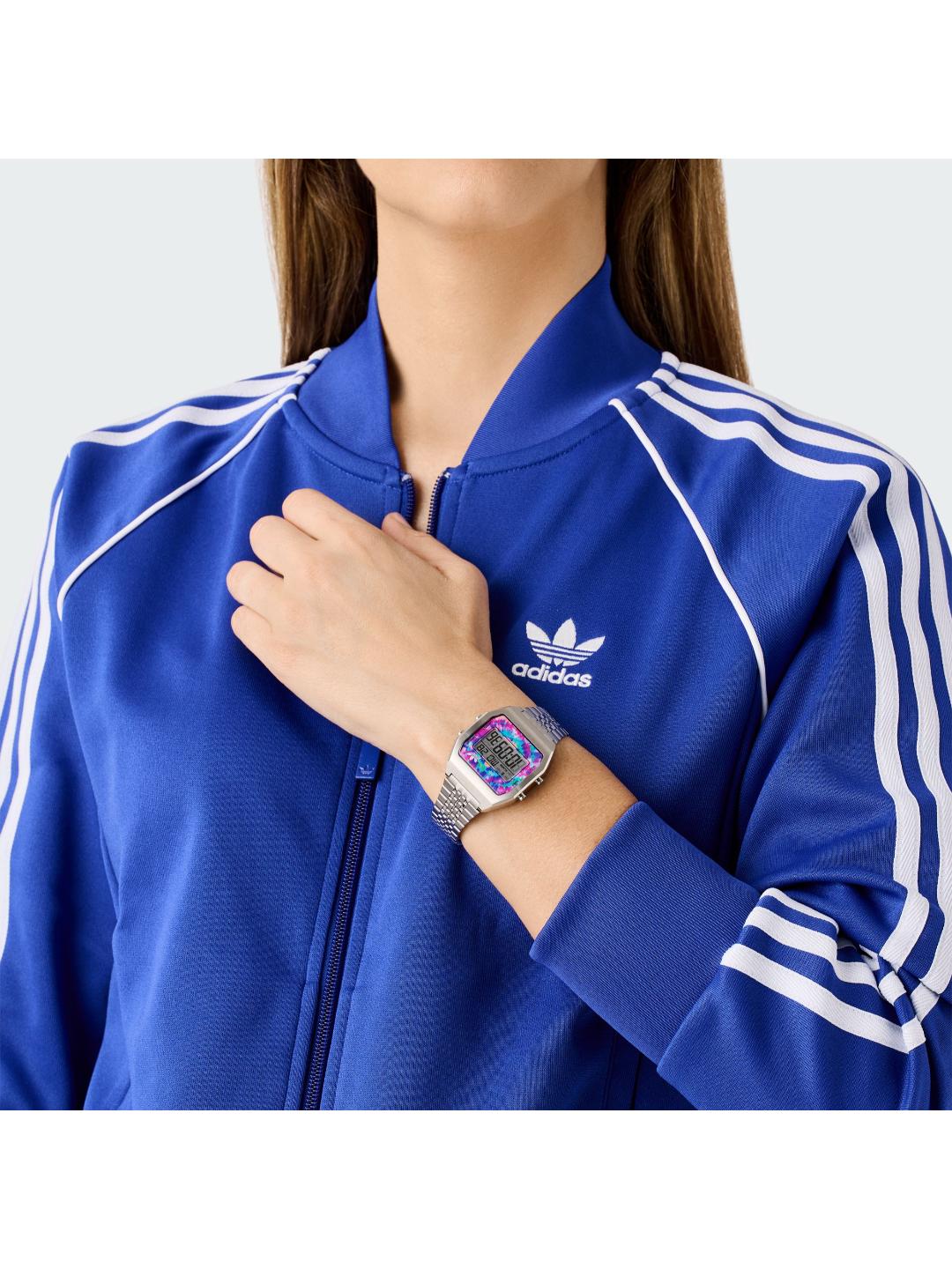 A close-up of a person wearing a royal blue Adidas track jacket with white stripes and a white Adidas logo on the chest, accessorized with a digital watch.