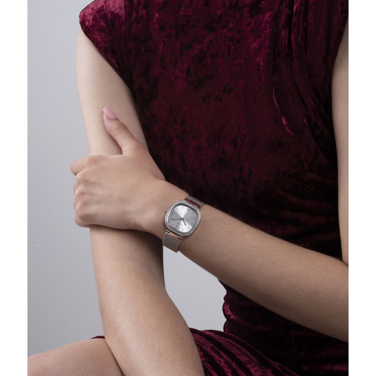 A silver-toned wristwatch with a square bezel and silver dial, worn on a model's wrist. The model is wearing a textured deep burgundy velvet dress against a plain light gray background.