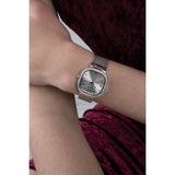 A silver-toned women's watch with a square bezel, crystal-embellished edges, and a mesh metal bracelet, shown worn on a wrist against a burgundy velvet background.