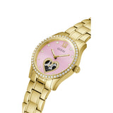 Gold-tone Guess women's watch with a pink dial, crystal-embellished bezel, and a heart-shaped skeleton window on the face, shown on a white background.