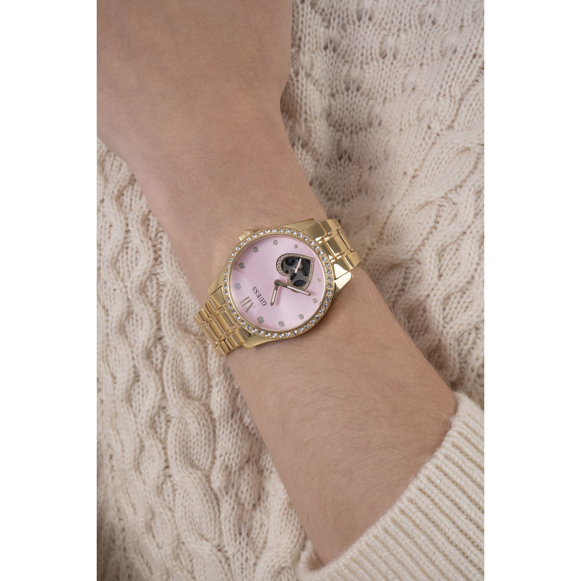 A gold-tone wrist watch with a light pink dial and crystal-studded bezel, shown worn on a person's wrist over a cream cable-knit sweater.