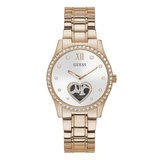 A Guess women's analog watch with a rose gold metal link bracelet, a white dial, a bezel set with clear rhinestones, and a heart-shaped crystal accent on the face.