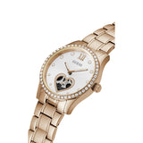 A Guess women's analog watch with a rose gold metal bracelet and case, featuring a white dial with Roman numeral XII, crystal rhinestone accents, and a heart-shaped skeleton dial detail.