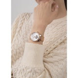A close-up of a woman wearing a rose gold wrist watch with a white dial and crystal-embellished bezel, paired with a cream-colored cable-knit sweater.
