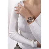 A close-up of a woman wearing a rose gold-toned watch with a metallic link bracelet and a patterned dial, paired with a white ribbed long-sleeve top.