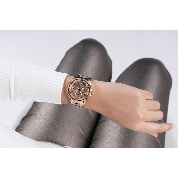 A rose gold-toned metal watch with a multi-dial face is shown on a woman's wrist, with her legs crossed in gray knit pants against a clean white background.