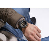 A close-up of a person's wrist wearing a black and gold chronograph watch, with blue jeans and a brown jacket visible in the background.