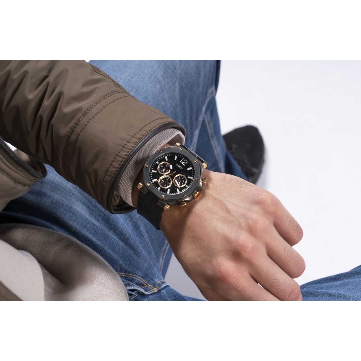 A close-up of a person's wrist wearing a black and gold chronograph watch, with blue jeans and a brown jacket visible in the background.
