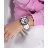 A wristwatch with a white strap, gold-tone case, and a crystal-studded bezel, shown worn on a person's wrist. The person is wearing a pink sweatshirt and white pants.