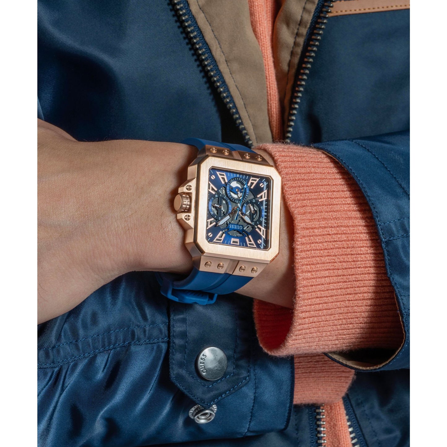 A close-up of a wrist wearing a rectangular rose gold watch with a blue dial and blue strap, worn under a blue jacket and peach ribbed sweater cuff.