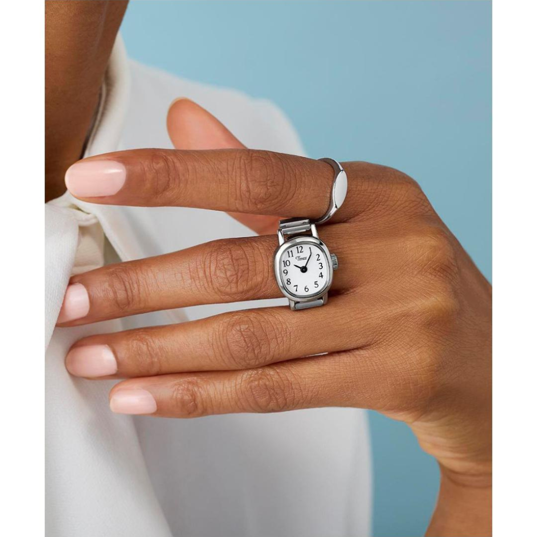 A silver-toned ring designed like a wristwatch with a round white face and black numerals, worn on a person's finger. The person is also wearing a slim silver band ring on another finger, against a light blue background.