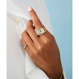 A close-up of a hand wearing a small gold watch with a white dial and a slim gold band, styled over a white shirt against a light blue background.