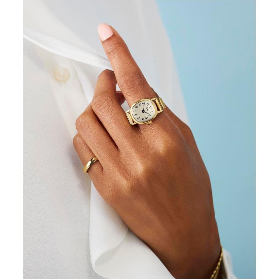 A close-up of a hand wearing a small gold watch with a white dial and a slim gold band, styled over a white shirt against a light blue background.