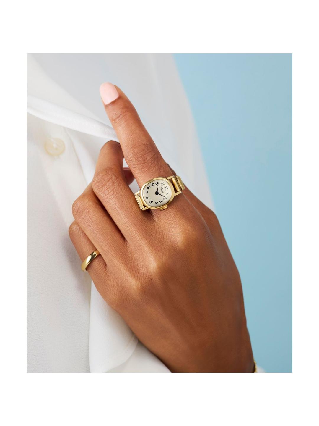 A close-up of a person's hand wearing a gold watch with a white dial and a gold ring on another finger, styled with a white shirt against a light blue background.
