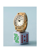 A gold-tone wristwatch with a white dial and black numerals, displayed on top of a white dice block featuring green and red playing card markings, against a pale blue background.